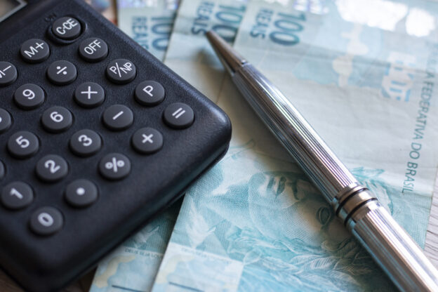 Calculadora, caneta e notas de cem reais representando a política fiscal e o controle econômico do governo.